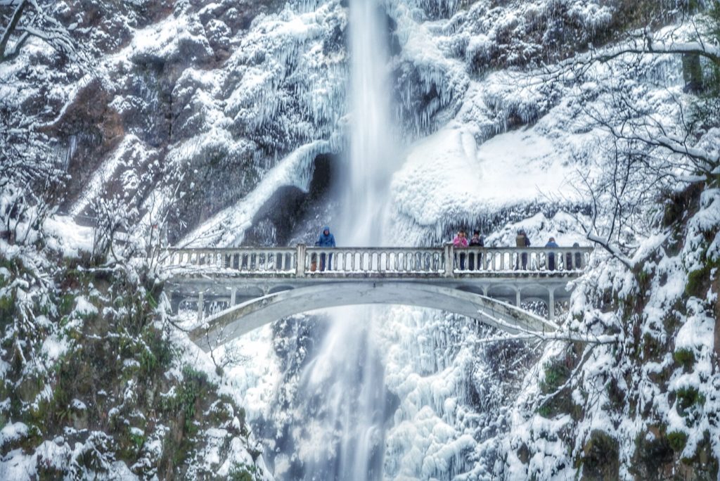 a photo of the bridge over Multnomah Falls in the winter, ice and snow covering the rocks and mosses and tree branches. people dressed in winter clothing are standing on the bridge