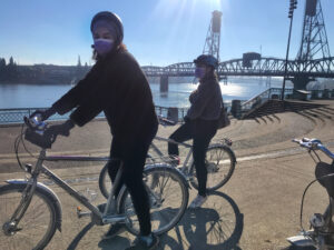 two young Black women on silver bikes smile behind their masks on a sunny day, Hawthorne Bridge in the background and Willamette River shining in the sun
