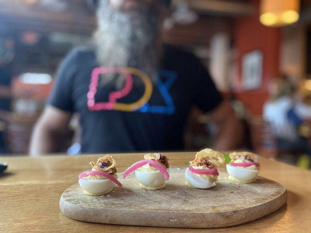 an oblong wooden tray of four deviled eggs, each balanced on a little pile of mustard-colored egg yolk puree, and each topped with pickled red onions, crispy shallots, and some bacon-like crumble, sits on a wooden table in front of a person with a t-shirt reading "PDX" in bold pink, yellow and blue font.