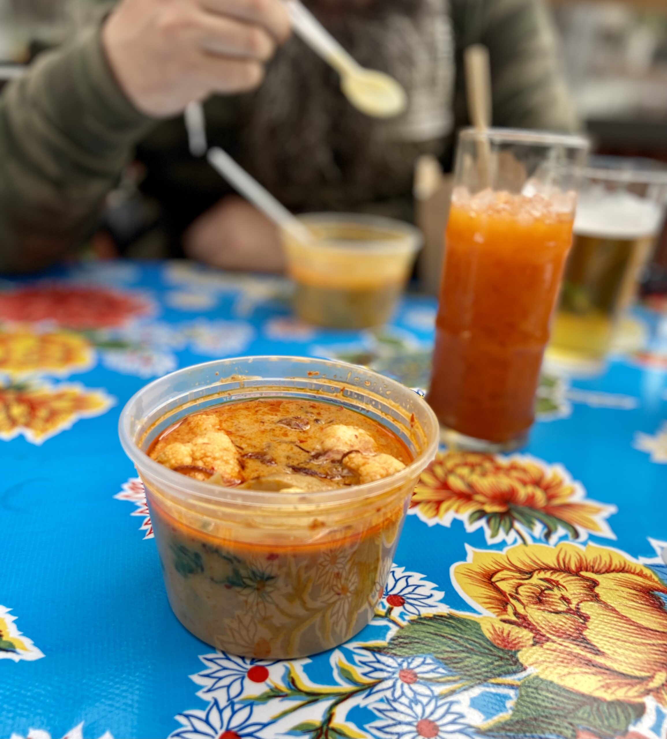 on a brilliant blue tablecloth decorates with yellow, red and blue flowers sits a plastic container of amber-colored curry, cauliflower and oyster mushroom pieces visible in the broth. behind the curry is a tall glass with an orange-amber colored beverage, behind that a pale amber beer and another darker curry container. a person with a camo sweatshirt is eating the curry