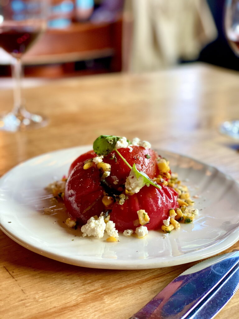beautiful crimson globes of heirloom tomatoes mounded on a white plate with brilliant yellow corn kernels, nuggets of soft cheese, and a coating of vinaigrette and green herbs. an out-of-focus glass of red wine is in the background