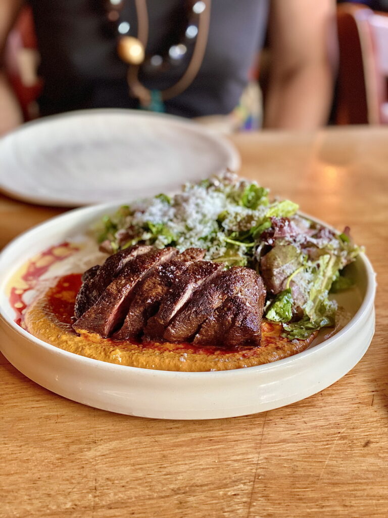 a round and shallow white bowl-style plate holds a bavette steak, cooked medium rare and balanced in a pale amber sauce, with a pile of fresh greens covered in vinaigrette, with a small white plate and a person wearing a chunky necklace behind