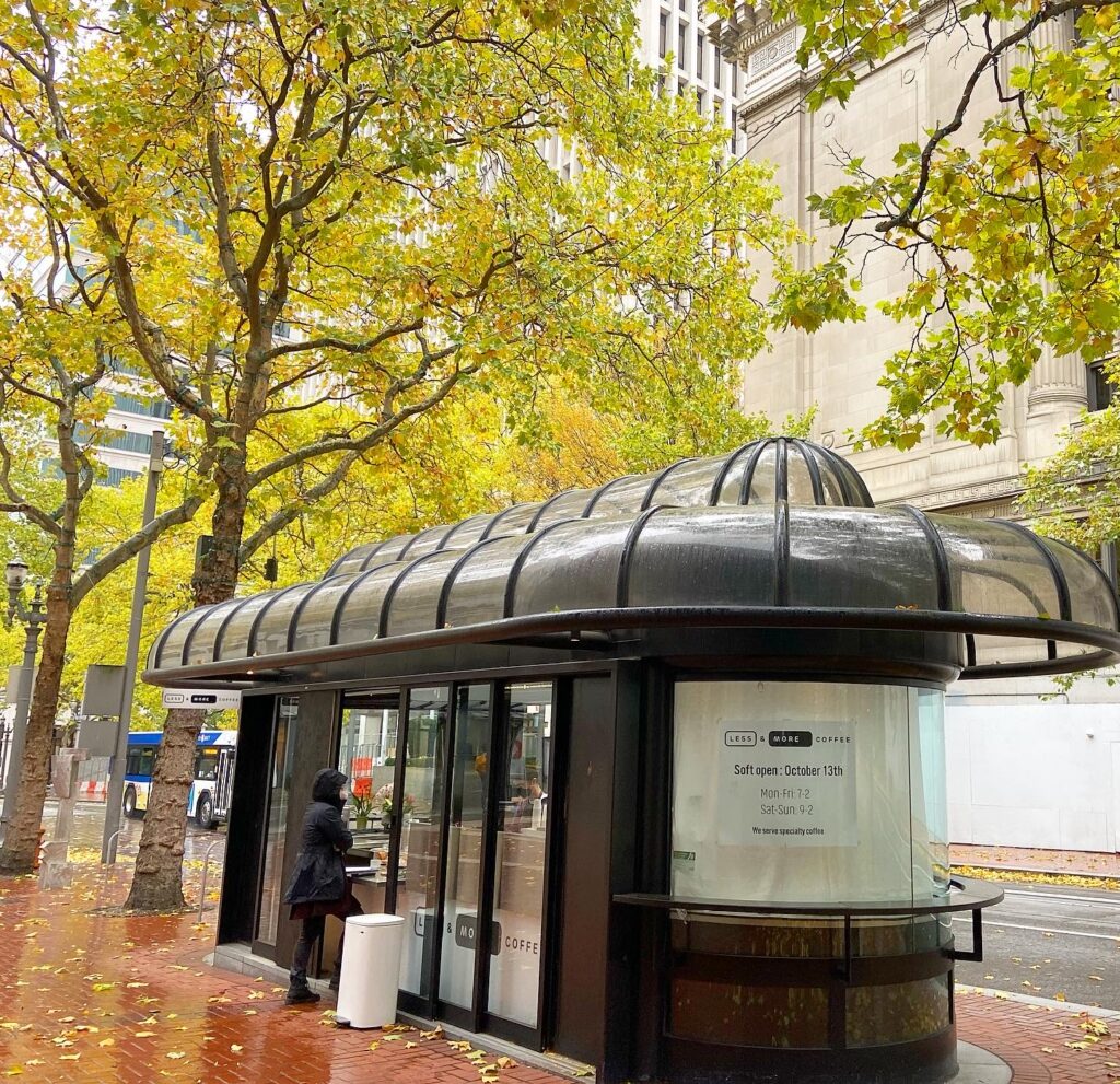 A 50s-style bus stop converted to a modern, minimalist coffee shop with a sign, “less and more coffee soft open october 13.” and “mon-fri 7-2, sat-sun 9-2.” A person wearing black rain gear stands in front ordering coffee. The sidewalk is soaked in rain, making the red brick luminous, and the trees surrounding the coffee shop are yellows and chartreuses. A city bus passes on the street behind.