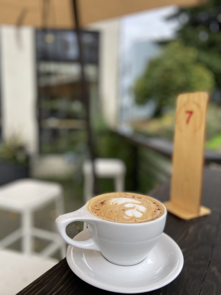 in a white cup on a white saucer, a beautiful amber colored latte sprinkled with spice sits on a dark brown table, a wooden table number with a red 7 on it in the background. you can see white stools and an amber colored umbrella over what seems to be an outdoor patio in the background. there is a white and dark brown coffee shop window out of focus behind