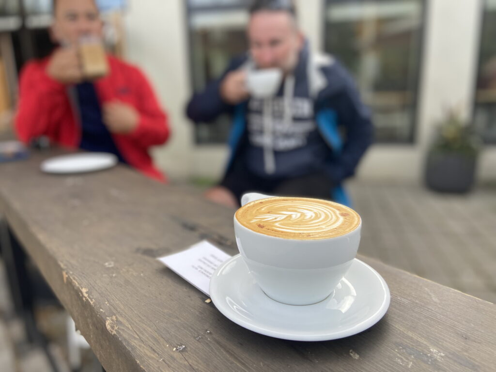 a beautifully made cappuccino with a circular floral latte art on the top sits in a white cup on a white saucer on a wooden bar-style table diagonally stretching into the frame. behind it are two people wearing blue and red jackets, each drinking a coffee drink, with the big black-framed windows in a white building that appear to be a coffee shop, behind. this is upper left roasters in Portland oregon