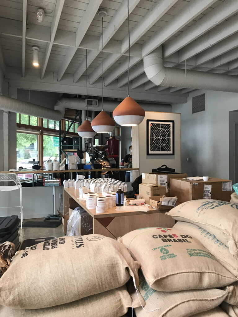 at Upper Left Roasters, burlap bags of coffee beans reading "cafes do Brasil" and "certified fair trade" sit in front of a table filled with coffee bags and labels; behind those are the coffee roasting machines. an image on the wall shows a graphical black design with a red triangle in the upper left corner