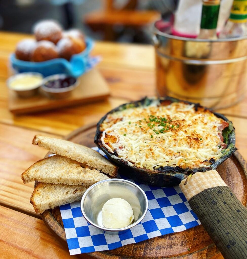 A skillet of eggs covered with spinach and cheese and breadcrumbs, browned in the center, skillet handle covered in a little Forest green hot pad and accompanied by three slices of toast and a ramekin of pale yellow butter on blue checked paper. Behind is a turquoise handled crock of round pancake balls sprinkled with powdered sugar and accompanied by ramekins of lemon curd and lingonberry jam. There is a silver bucket of condiments. Everything is on a picnic style table that appears to be outside.