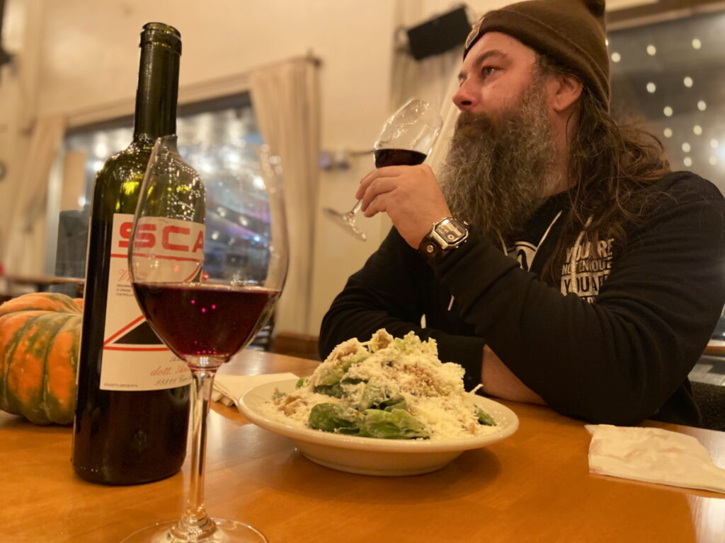 In dim evening lighting, a person with a big beard and a brown beanie holds a glass of red wine, white bowl of Caesar salad absolutely drowned in Asiago/Parmesan cheese and small bread crumbs in front of him on a wooden bar next to a glass of red wine and the bottle, which has the horizontal letters “SCA” in red. Behind is an orange and green striped squash and a numbered sign