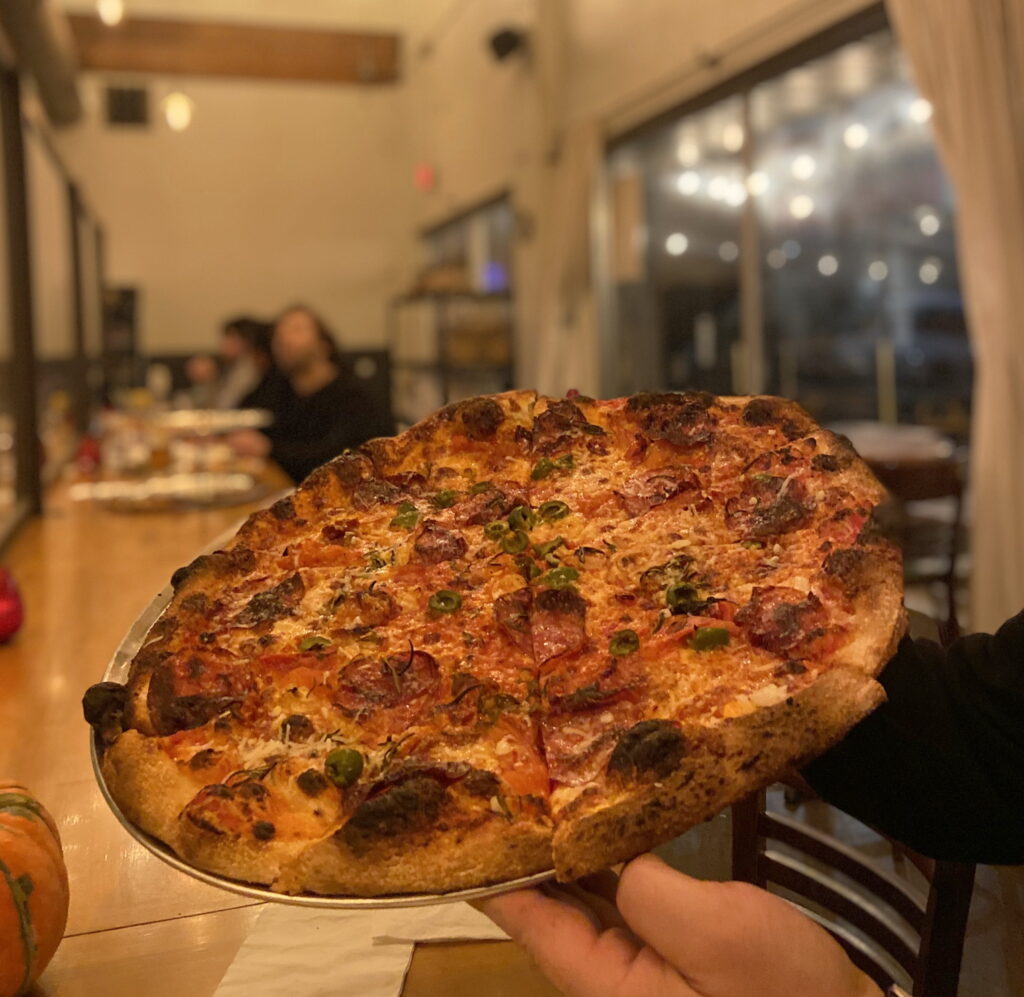 A large round pizza with charred and bubbly crush and a variety of toppings including soppressata and rosemary and cheeses, held in a hand in front of a bar with a pumpkin and other people behind