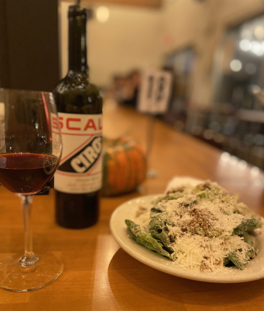 In dim evening lighting, a white bowl of Caesar salad absolutely drowned in Asiago/Parmesan cheese and small bread crumbs sits on a wooden bar next to a glass of red wine and the bottle, which has the horizontal letters “SCAL” in red and diagonal letters “CIRO” in white on a dark blue banner. Behind is an orange and green striped squash and a numbered sign 
