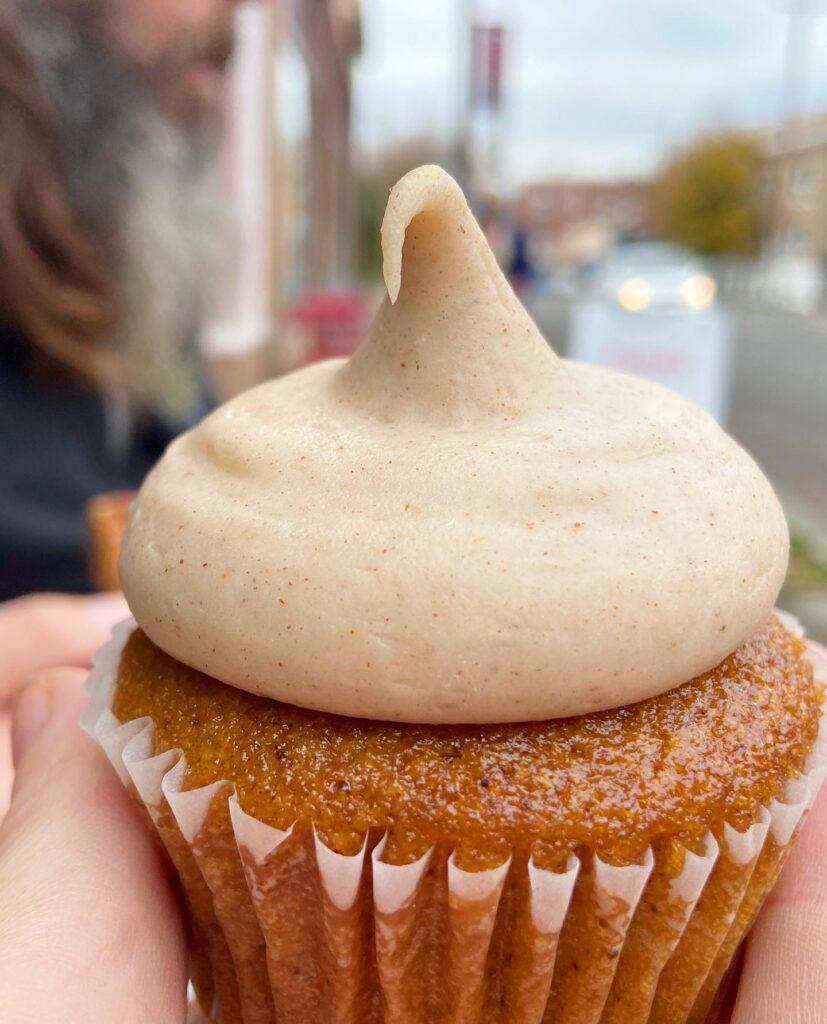 a pumpkin cupcake with a kiss-shaped dollop of pumpkin spiced cream cheese frosting held in a hand on a city street in the fall