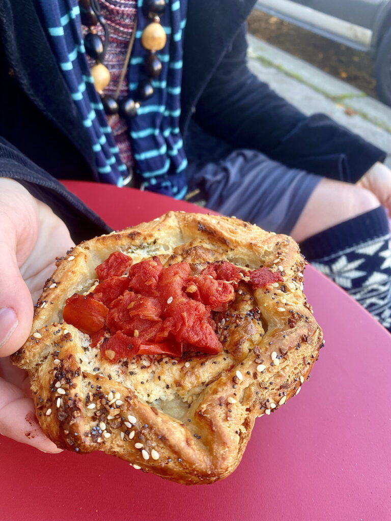 a person in dark striped clothes at a red table holding a round galette pastry topped with jewel-red tomato chunks and covered in sesame seeds, poppy seeds and onion bits. the crust looks very flaky.
