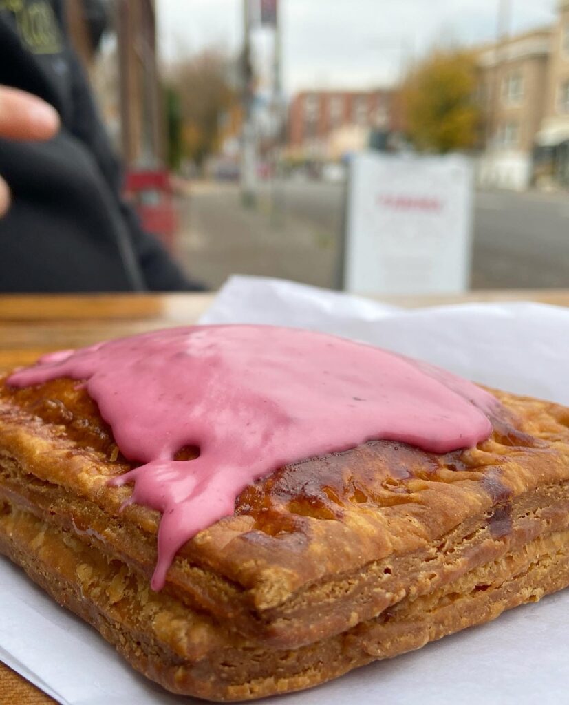 a rectangular flaky pastry topped with tantalizingly dripping pink frosting sits on a table with a hand reaching toward it. a fall scene on a city street is in the background, and you can just make out a white a frame sign that might say "farina" in red letters
