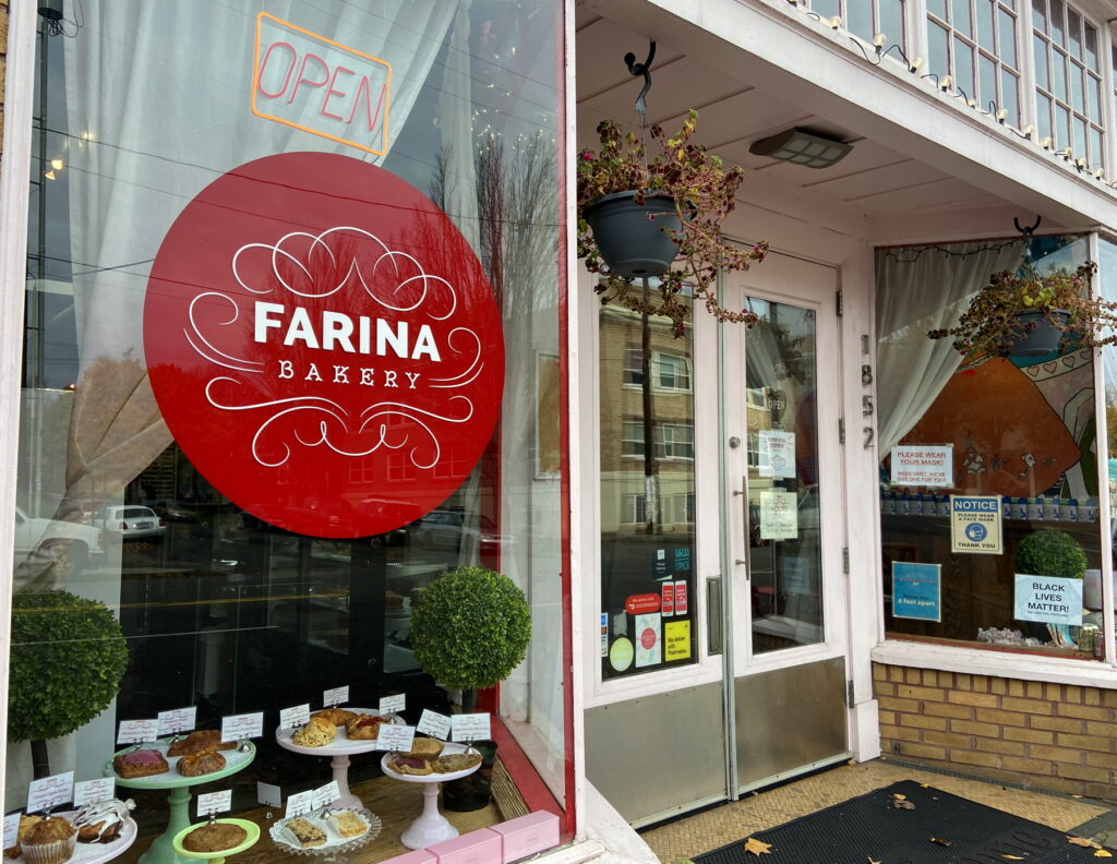 a pretty storefront of Farina Bakery, with a flourished red painted sign and a light pink paint around big windows and a windowed door. in the front window are a number of footed plates with baked goods and little tags describing them.