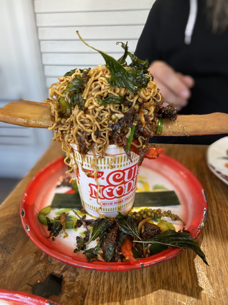a cup of noodles package on a red-rimmed plate with crispy basil leaves, bell pepper slices, and a green coriander seed pod spilling over onto the plate. on tope of the cup of noodles container is a mass of noodles, red spices, beef and more crispy basil leaves balanced on a bone. a person's black sweatshirt and hand are behind.