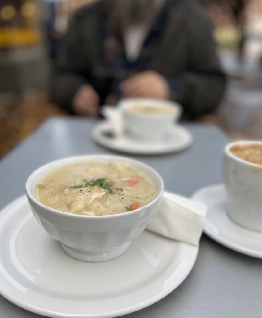 on a silver table on a street during a wet fall day are two white bowls on white plates, and a coffee cup on a white saucer. In the front bowl you can see a creamy chicken soup with carrots and sprinkled with green herbs and black pepper. A person in a dark coat sits behind the table.