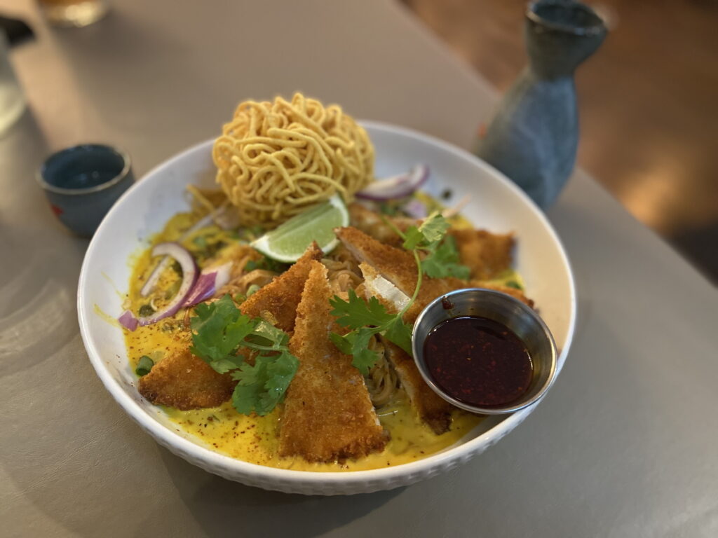 Photo of a dish of Thai food surrounded by a blue sake cup and carafe. In a yellow broth speckled with chile and red onions are wedges of katsu chicken, a ramekin of chile oil, and a lime wedge. In the back is a huge ball of fried udon. There are sprigs of cilantro over the top.