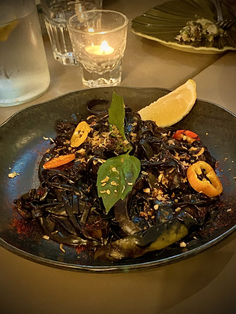 surrounded by water carafe, glowing votive candle, and water glass, is a black ceramic plate with even darker black linguine in a black sauce. on the side of the plate is a lemon wedge; sliced peppers and basil decorate the top. in the distance is a green leafy plate of Brussels sprouts