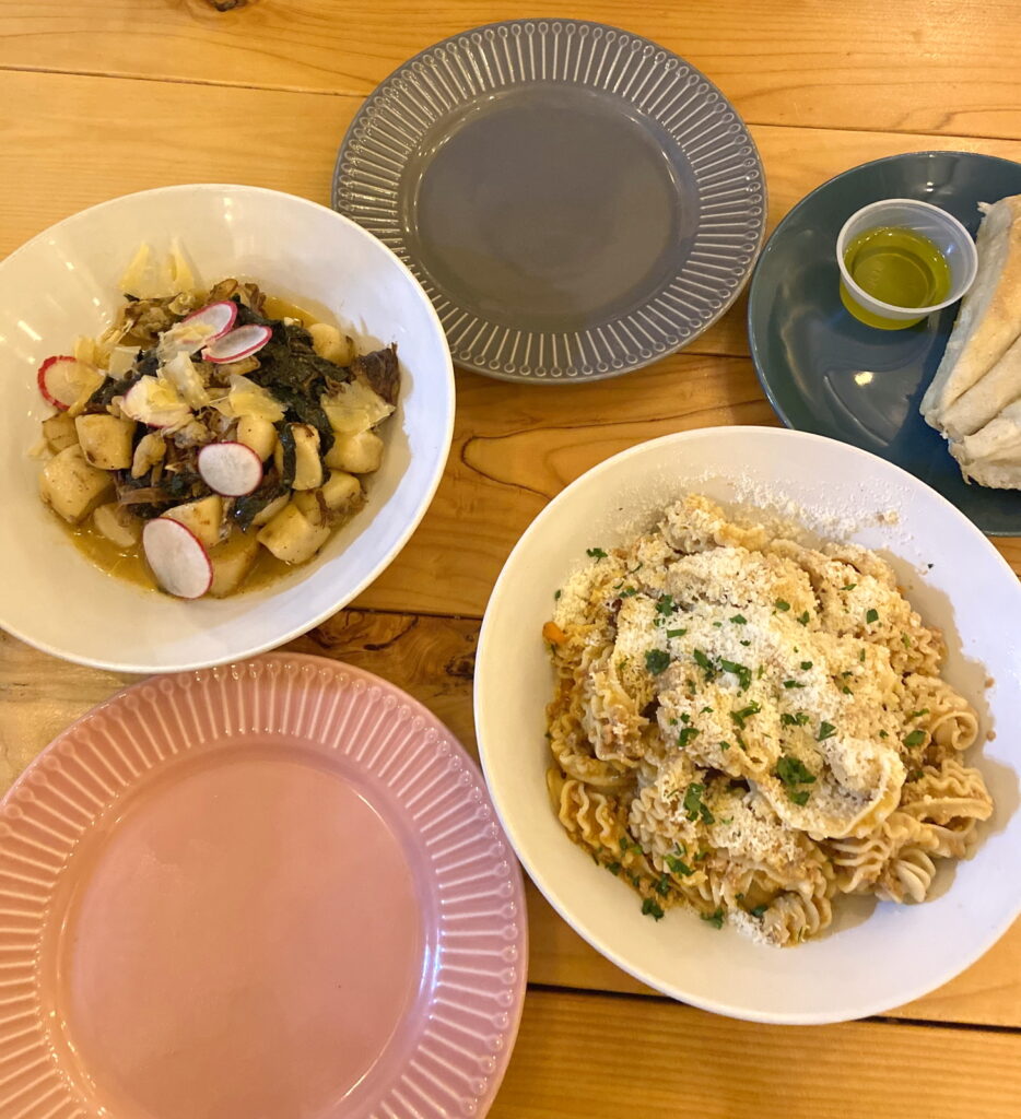 on a blond wood picnic style table, wide white bowls filled with radiatori style pasta and a root vegetable focused dish. the radiatori style pasta is covered with a bolognese sauce and parmesan and herbs. the root vegetable dish is topped with radish slices and some kind of green. there are colored plates; a pink and slate grey one are empty and a dark blue one has wedges of bread and a little container of olive oil.