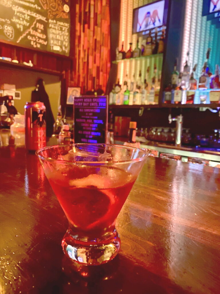 a red cocktail with red lighting behind it and a twist in it. There are lights behind