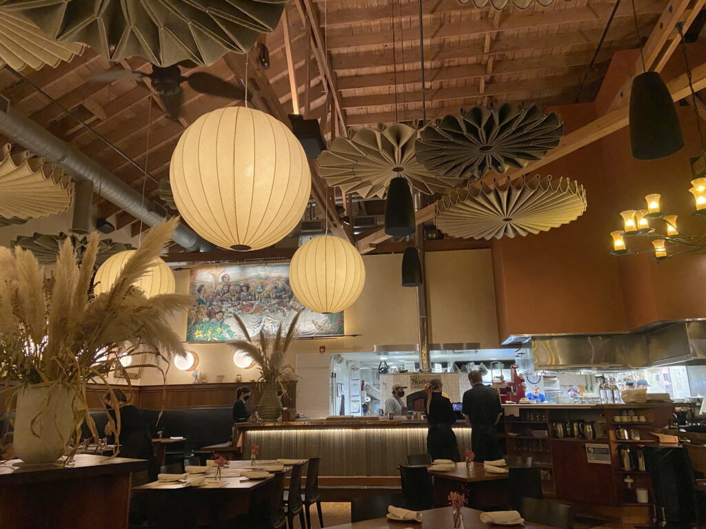 a stunning restaurant space with the kitchen at the back and beautiful soundproofing rosettes and large cylindrical paper lamps handing from the ceiling. people are working in the distance wearing black clothes and masks. there is a big vase of pampas grass in the foreground and many tables set with place settings