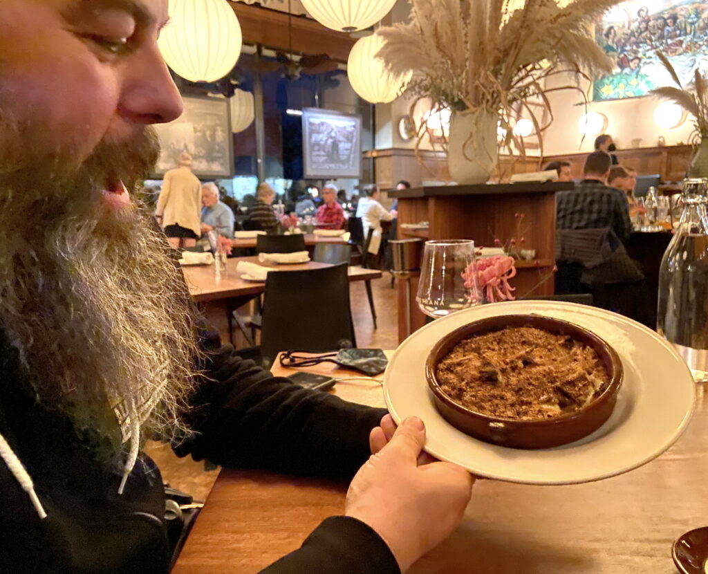 a person with a beard holds a dessert in a terra cotta crock on a white plate in a busy restaurant
