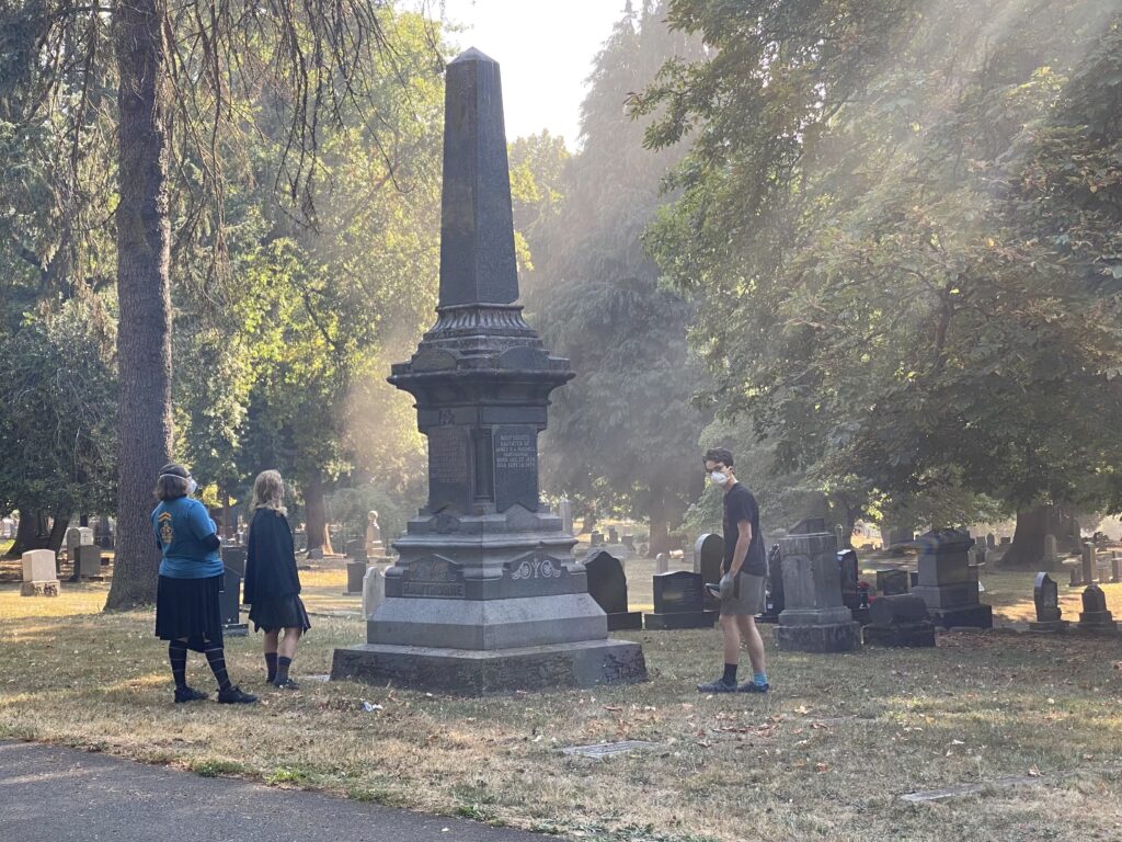 as sun cants through the haze, an obelisk is lit from behind and above. trees surround it. a few people are speaking in front. one wears a black cape and black skirt.