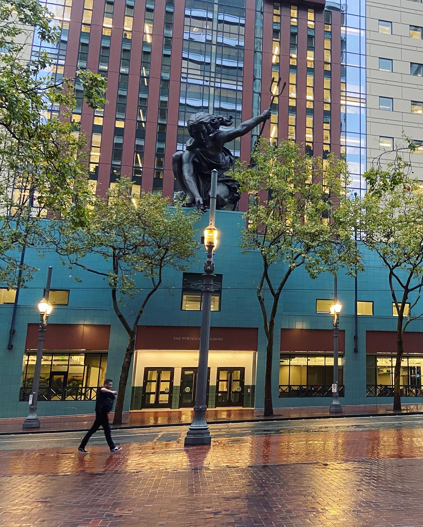 a rainy sidewalk reflects the lights and building above it, topped with a copper repousse figure holding a trident