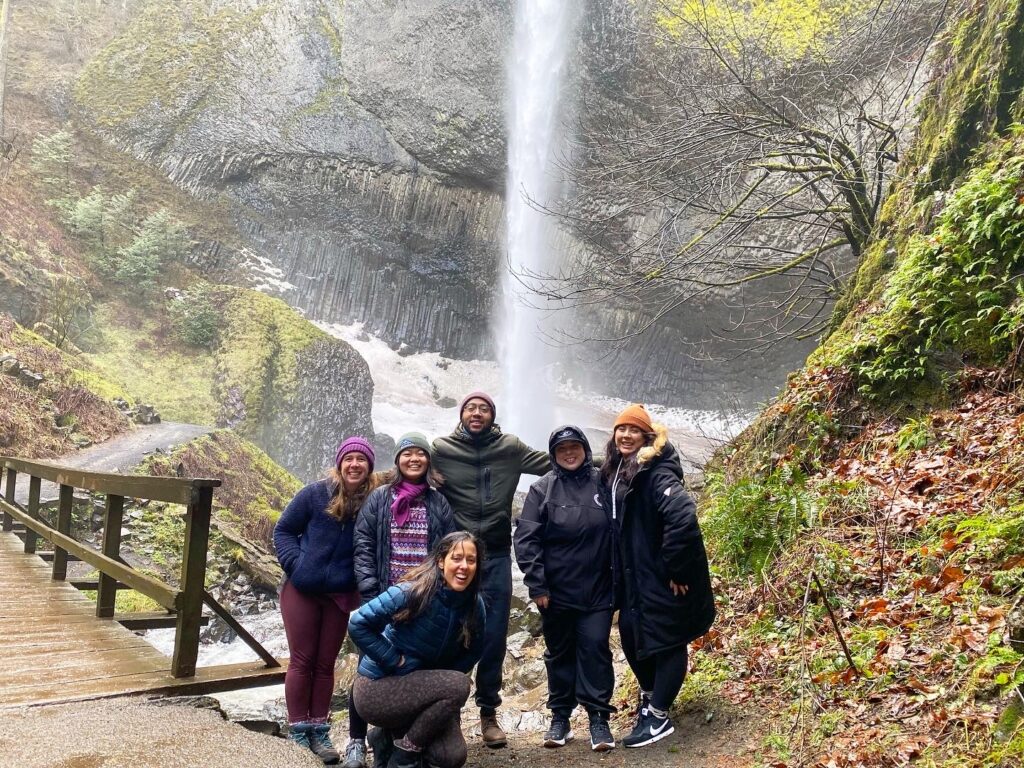 a group of people at the base of a waterfall, smiling. they are all wearing hats and hooded raincoats and look very cozy