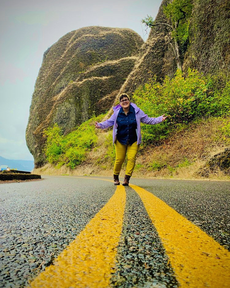 yellow lines on a road approach a person standing in the middle, with a bulbous mushroom shaped rock behind. the person is smiling and wearing mustard yellow pants and a purple hooded jacket and holding their arms wide