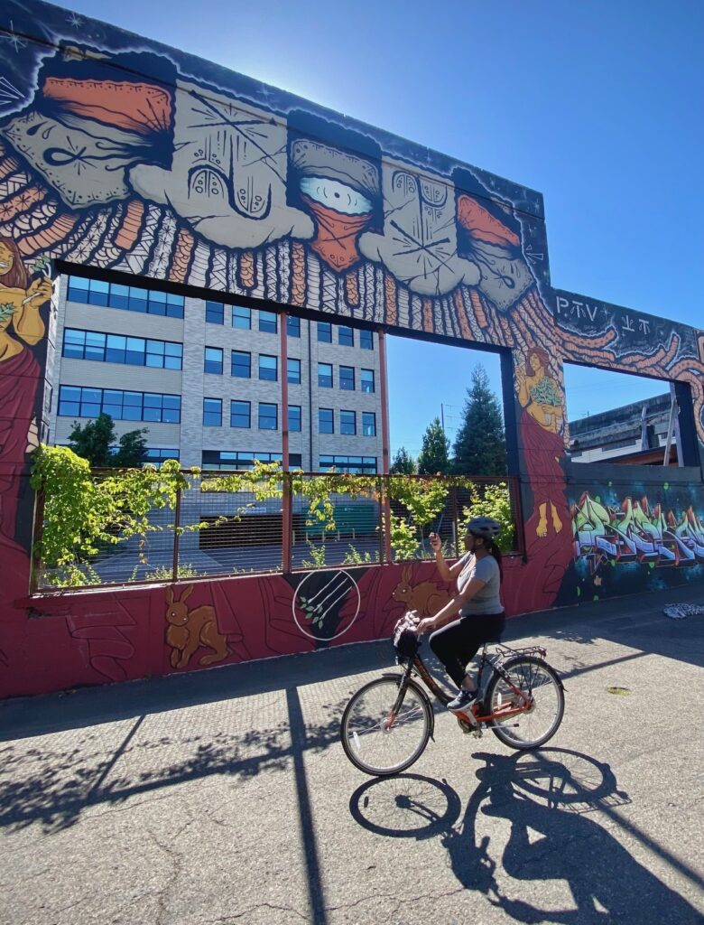 a person bikes past a big window-shaped concrete wall festooned with art on a sunny day